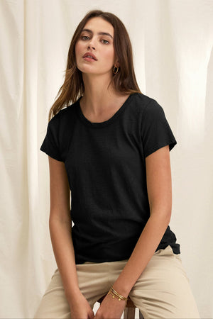 A person sits against a light fabric backdrop, wearing a TILLY TEE by Velvet by Graham & Spencer and beige pants, exuding the relaxed vibe of a California classic with their neutral expression.