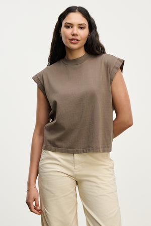 A woman with long dark hair wears the ASYA TEE by Velvet by Graham & Spencer, a vintage-inspired cropped muscle tee in soft cotton jersey, paired with beige pants against a plain light background.