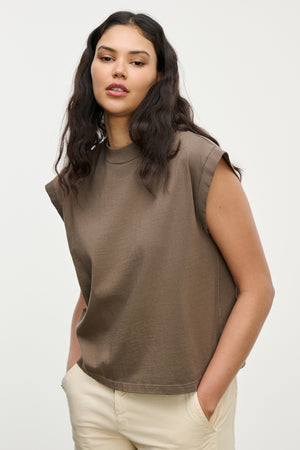 A woman wears the Velvet by Graham & Spencer ASYA TEE, a vintage-inspired sleeveless brown cropped muscle tee, paired with beige pants and standing hands-in-pockets against a plain light background.