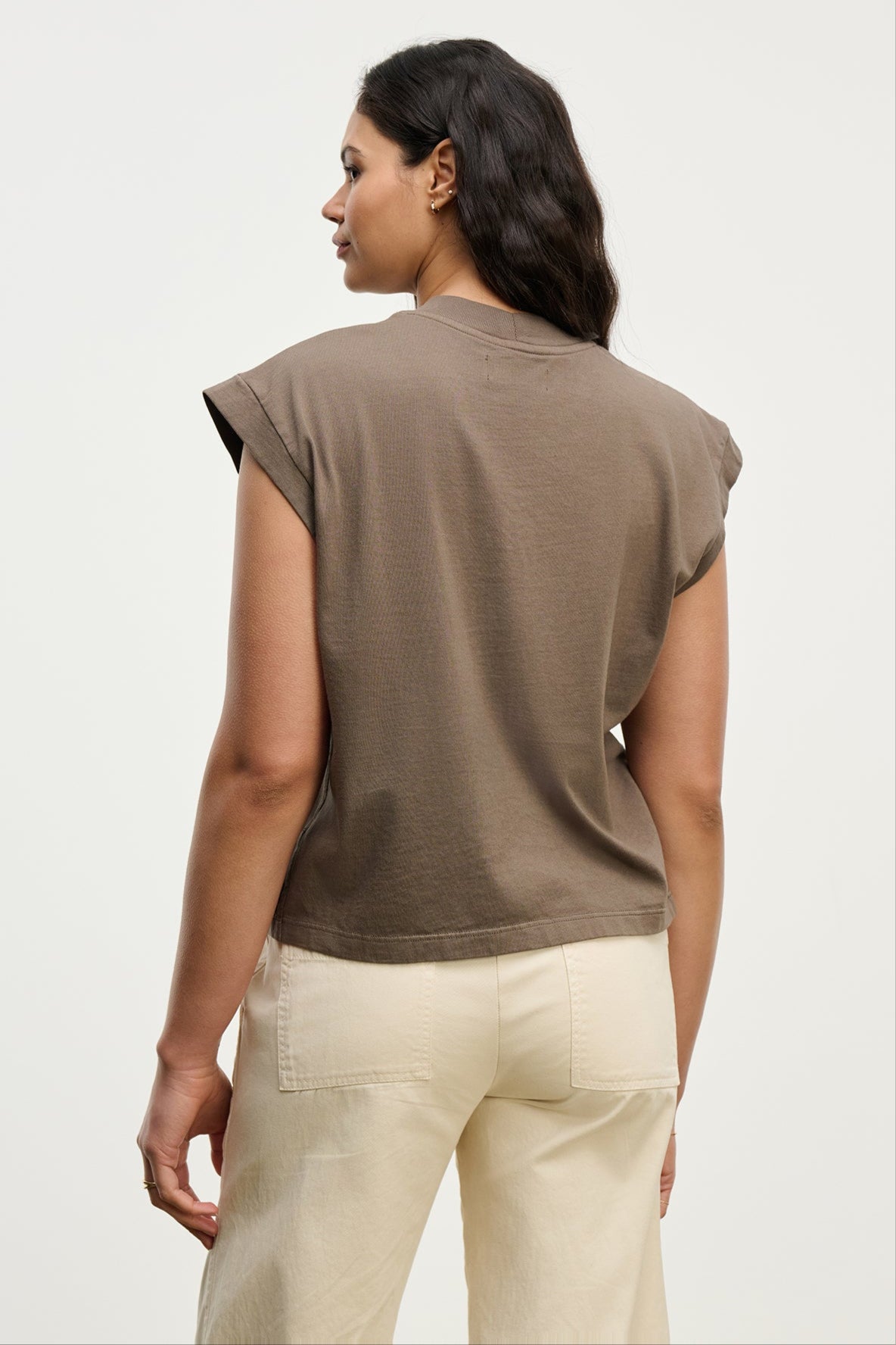 A woman faces away from the camera, wearing the vintage-inspired brown ASYA TEE by Velvet by Graham & Spencer and cream pants against a plain light background.
