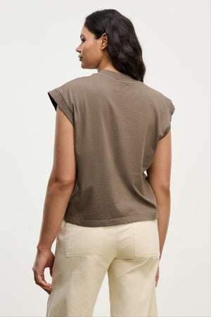 A woman faces away from the camera, wearing the vintage-inspired brown ASYA TEE by Velvet by Graham & Spencer and cream pants against a plain light background.