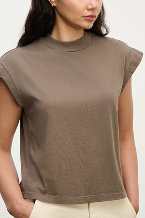 A woman wearing the ASYA TEE by Velvet by Graham & Spencer, a vintage-inspired brown cotton jersey crew neck shirt with cuffed short sleeves, pairs it with cream-colored pants against a plain background.