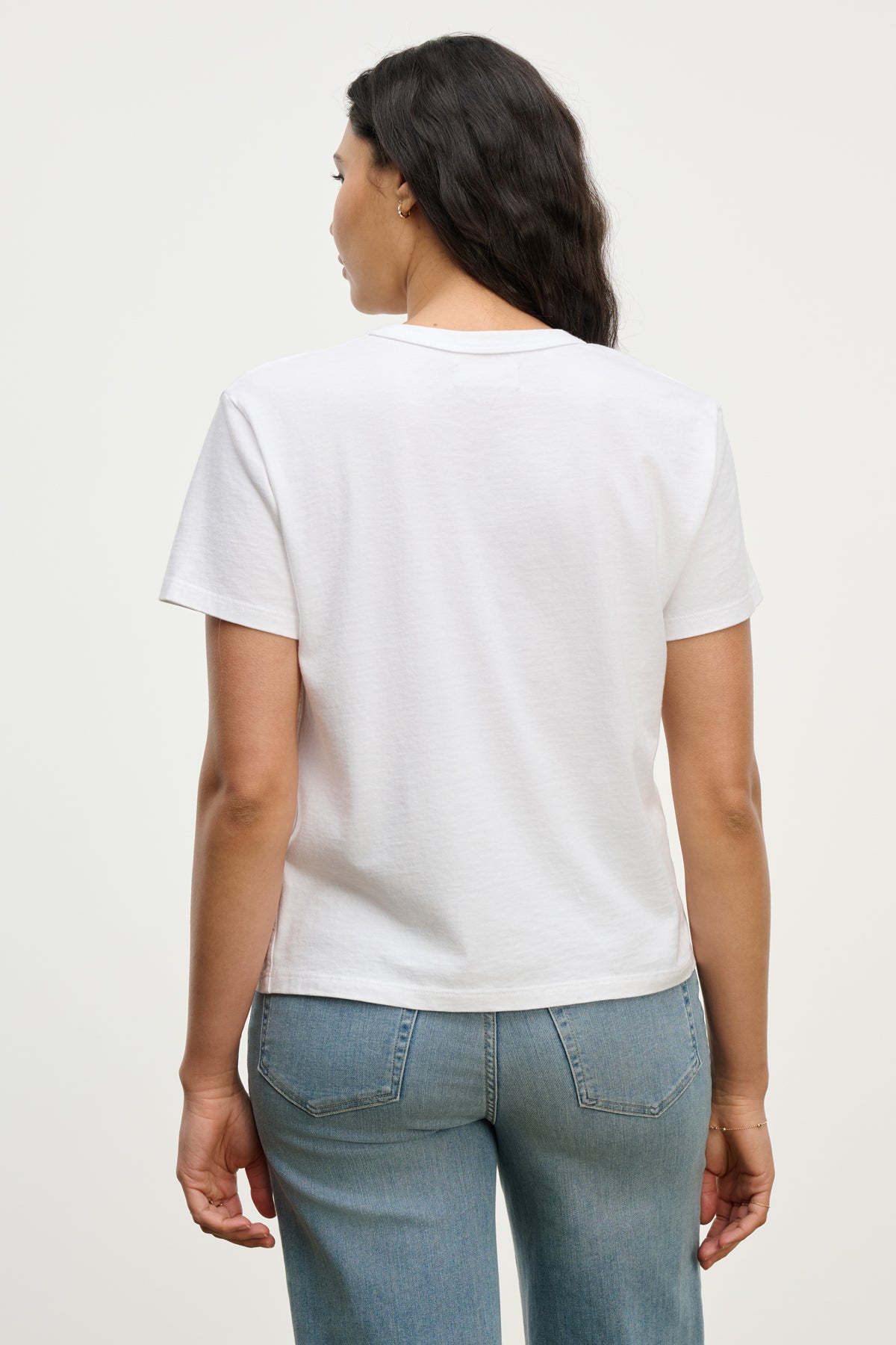 A woman faces away from the camera, wearing the LEIGH TEE by Velvet by Graham & Spencer, a vintage-inspired soft cotton jersey, paired with light blue jeans against a neutral background.-39742241603777