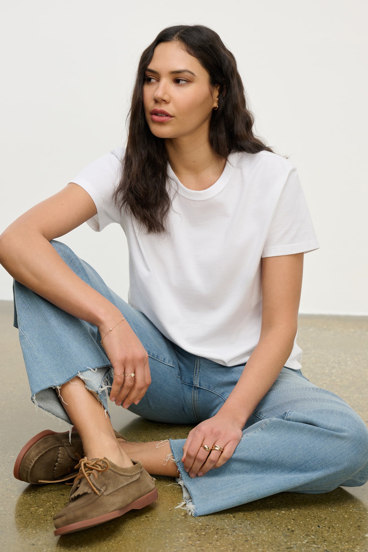 A woman with long dark hair wears the LEIGH TEE by Velvet by Graham & Spencer, styled with light blue jeans and brown shoes, as she sits on the floor against a light background.-39742241538241
