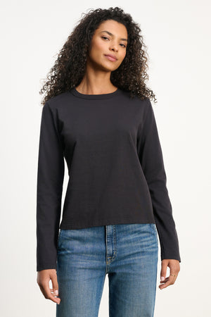 A woman with curly hair wears the SUSANNA TEE by Velvet by Graham & Spencer, a vintage-inspired long-sleeve black cotton jersey shirt, paired with blue jeans against a white background.