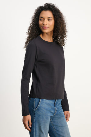A woman with curly hair wears the SUSANNA TEE by Velvet by Graham & Spencer—a vintage-inspired, long-sleeved black cotton jersey shirt—paired with blue jeans, standing against a plain white background and looking slightly left.
