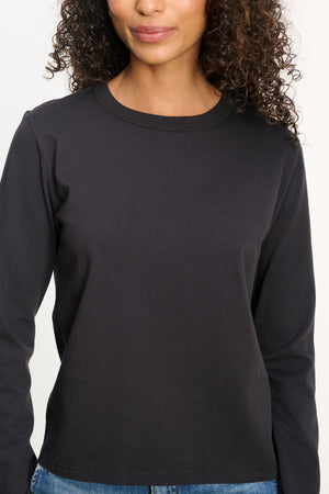 A woman with curly hair wears the SUSANNA TEE from Velvet by Graham & Spencer—a vintage-inspired, long-sleeve black cotton jersey top—paired with blue jeans, shown from shoulders to hips against a plain background.
