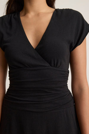 A woman models the ELLA DRESS by Velvet by Graham & Spencer, a black V-neck short-sleeve wrap dress in soft cotton jersey, shown from shoulders to waist against a plain background for a flattering look.