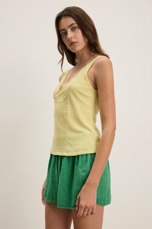 A woman stands against a plain background, wearing the HANNAH LACE TANK TOP by Velvet by Graham & Spencer in yellow, featuring soft cotton fabric with lace detailing, paired with green shorts.
