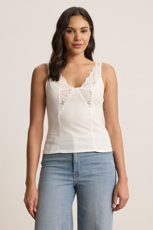 A woman stands against a plain background wearing the HANNAH LACE TANK TOP by Velvet by Graham & Spencer—a soft cotton white tank with lace trim along the V-neckline—paired with light blue jeans.