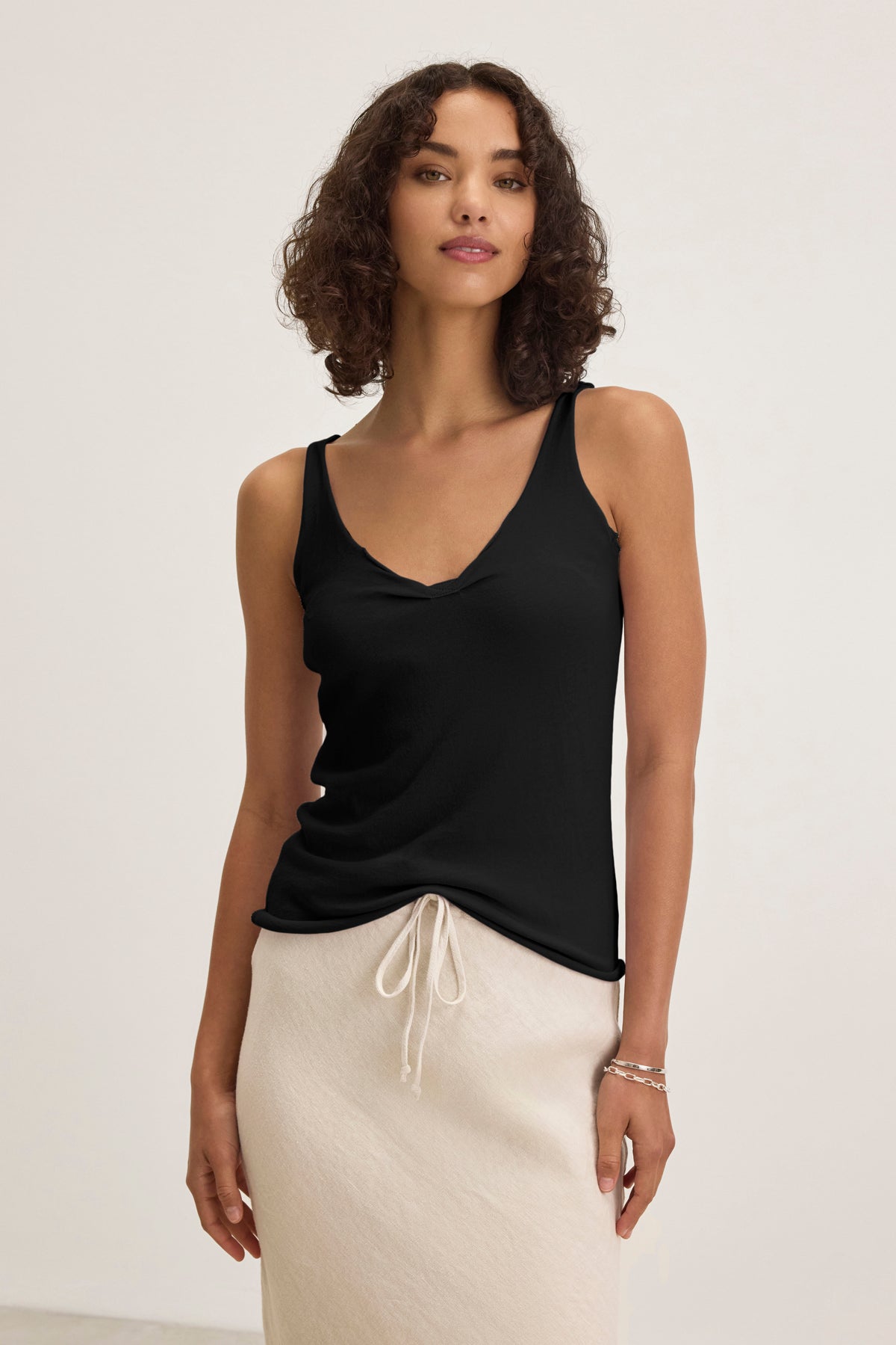 A woman poses against a plain background, wearing the KIM TANK TOP by Velvet by Graham & Spencer with a scoop neckline and a light beige drawstring skirt—a versatile wardrobe essential.-39173136253121