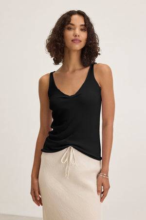 A woman poses against a plain background, wearing the KIM TANK TOP by Velvet by Graham & Spencer with a scoop neckline and a light beige drawstring skirt—a versatile wardrobe essential.