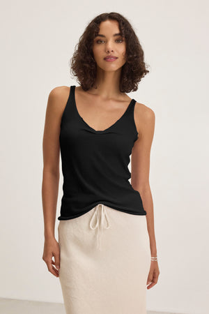 A woman stands against a plain light background, wearing the Velvet by Graham & Spencer KIM TANK TOP in black with a scoop neckline, paired with a beige skirt featuring a drawstring waist—a versatile wardrobe essential.
