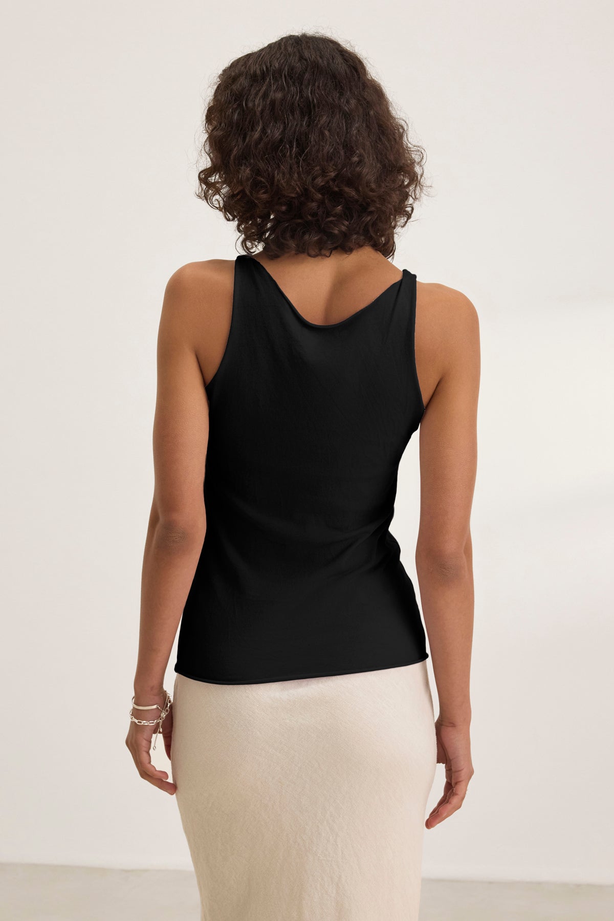 A woman with curly hair faces away from the camera, wearing the Velvet by Graham & Spencer KIM TANK TOP in black with a scoop neckline and no sleeves, paired with a light skirt against a plain background.-39173136318657