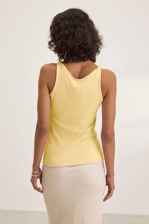 A woman with curly hair stands facing away in a well-lit room, wearing the KIM TANK TOP by Velvet by Graham & Spencer—a cotton tank with a scoop neckline—paired with a light skirt, showcasing timeless wardrobe essentials.