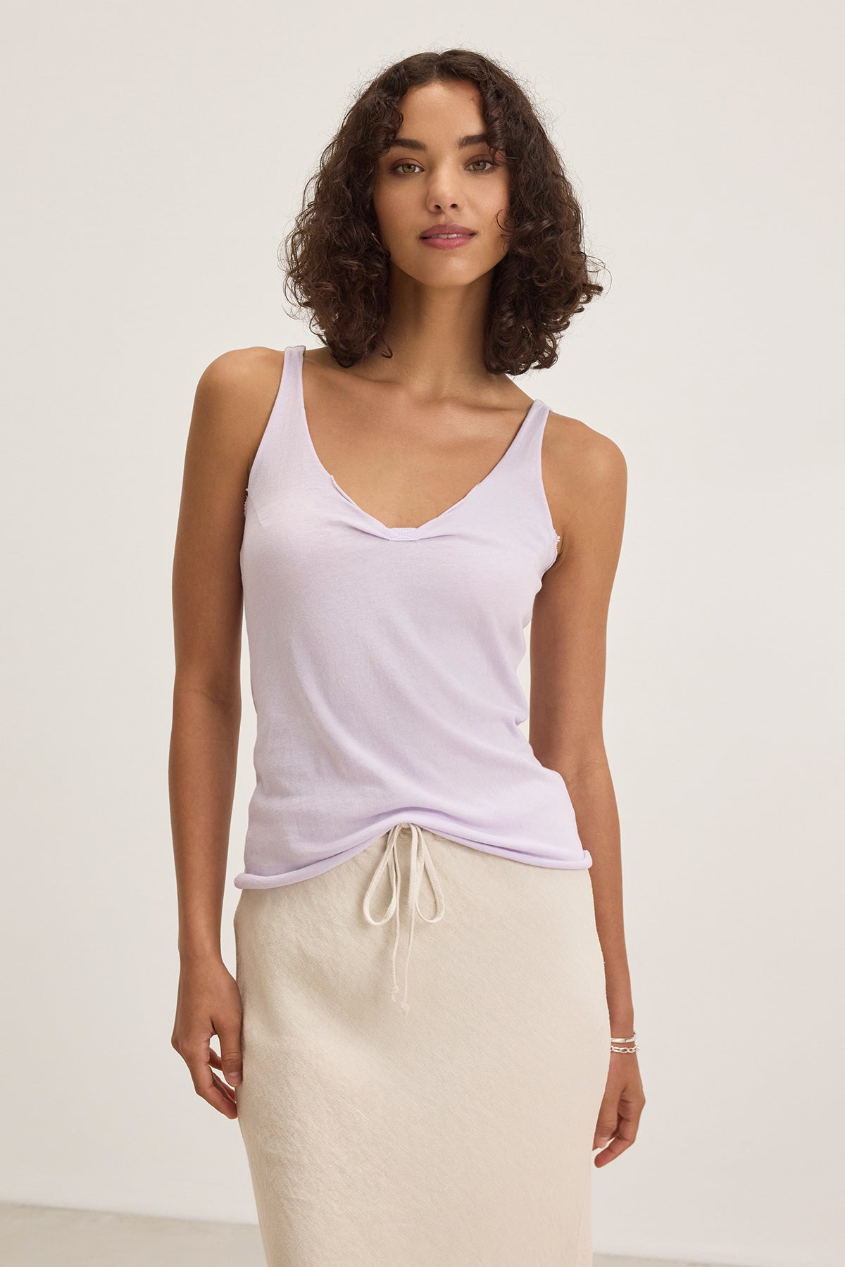 A woman in Velvet by Graham & Spencer's KIM TANK TOP, a lightweight cotton tank in light purple with a scoop neckline, pairs it with a cream skirt against a white background. This versatile essential is perfect for any occasion.