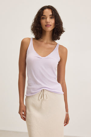 A woman in Velvet by Graham & Spencer's KIM TANK TOP, a lightweight cotton tank in light purple with a scoop neckline, pairs it with a cream skirt against a white background. This versatile essential is perfect for any occasion.