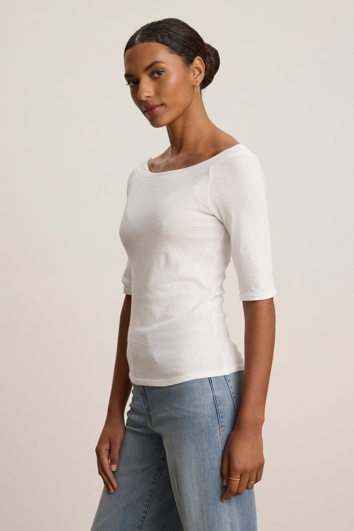 A woman stands sideways in the LUCILLE TEE by Velvet by Graham & Spencer, a white cotton jersey boat neck top with three-quarter sleeves, paired with light blue jeans for a casual look against a neutral background.-39148727042241