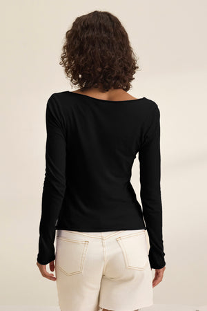 A person with curly brown hair is seen from the back wearing the Velvet by Graham & Spencer NOURA TEE, a classic black long-sleeve cotton jersey top, paired with white shorts against a plain light background.