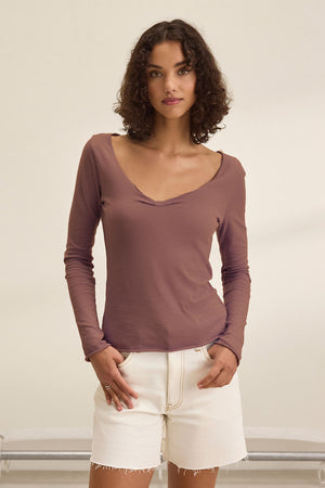 A woman with curly hair models the Velvet by Graham & Spencer NOURA TEE, a soft cotton jersey long-sleeve mauve top, paired with cream shorts against a plain light background.