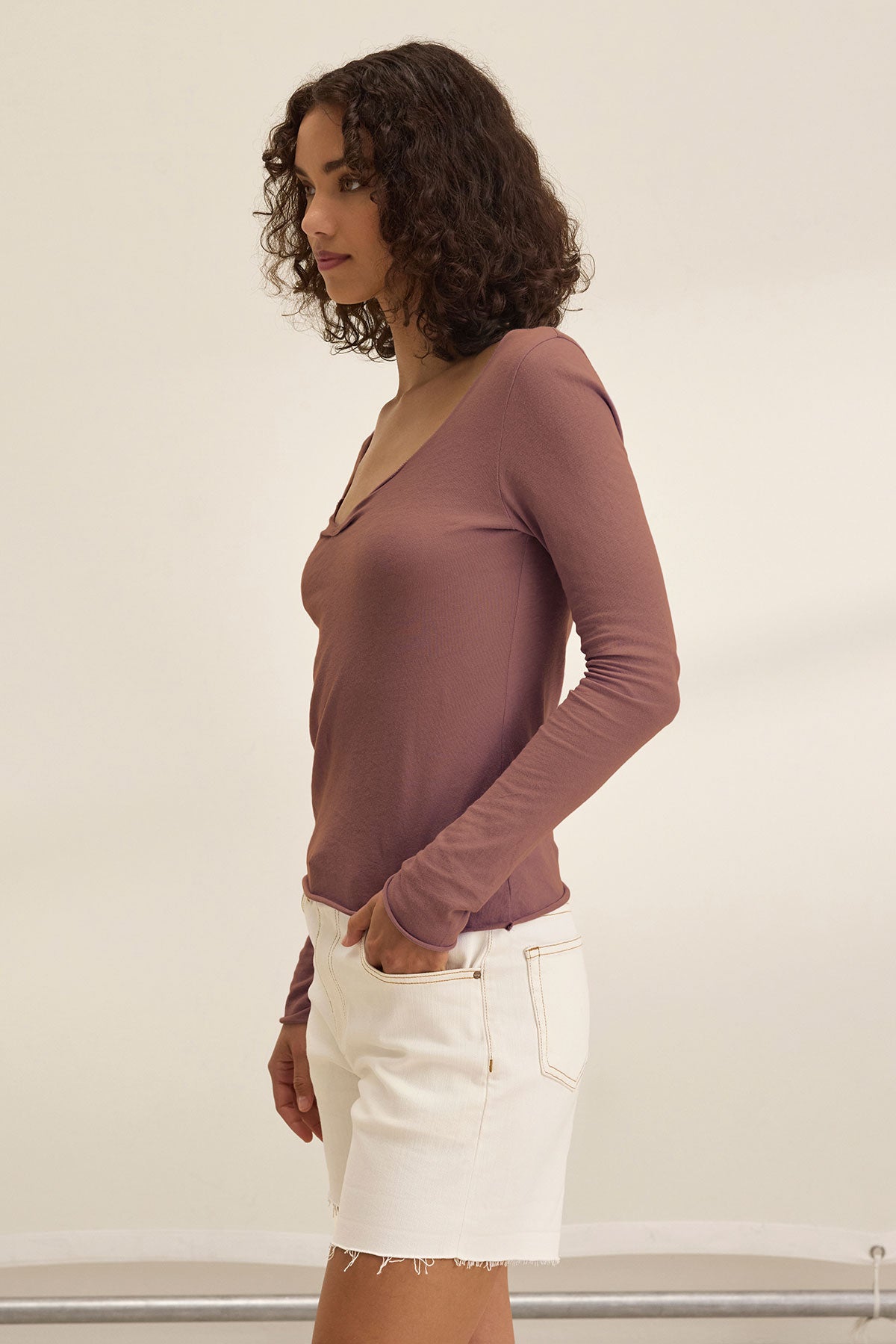 A woman with curly hair stands in profile against a plain background, wearing the mauve NOURA TEE by Velvet by Graham & Spencer paired with white denim shorts.