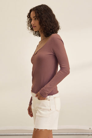 A woman with curly hair stands in profile against a plain background, wearing the mauve NOURA TEE by Velvet by Graham & Spencer paired with white denim shorts.