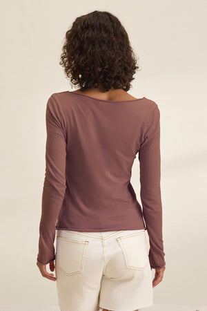 A person with curly hair is shown from the back, wearing the NOURA TEE by Velvet by Graham & Spencer—a long-sleeved mauve scoop neck shirt—paired with white shorts.