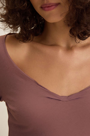 A woman with curly brown hair wears the NOURA TEE by Velvet by Graham & Spencer, a mauve V-neck classic style in soft cotton jersey; the image is cropped to her lower face, neck, and upper chest.