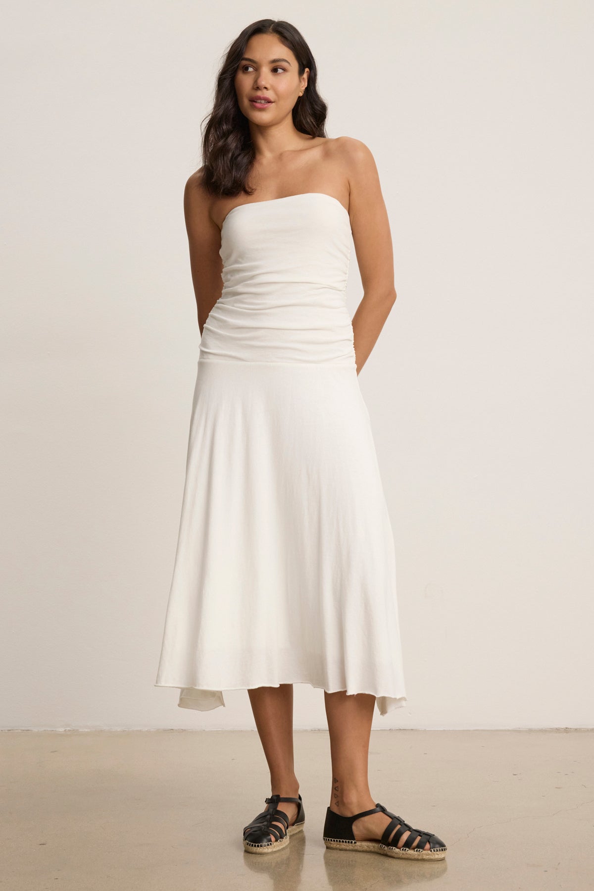 A woman stands indoors with her arms behind her back, wearing the strapless SONIA DRESS by Velvet by Graham & Spencer that creates a fitted, feminine silhouette, paired with black espadrille sandals.-39145533276353