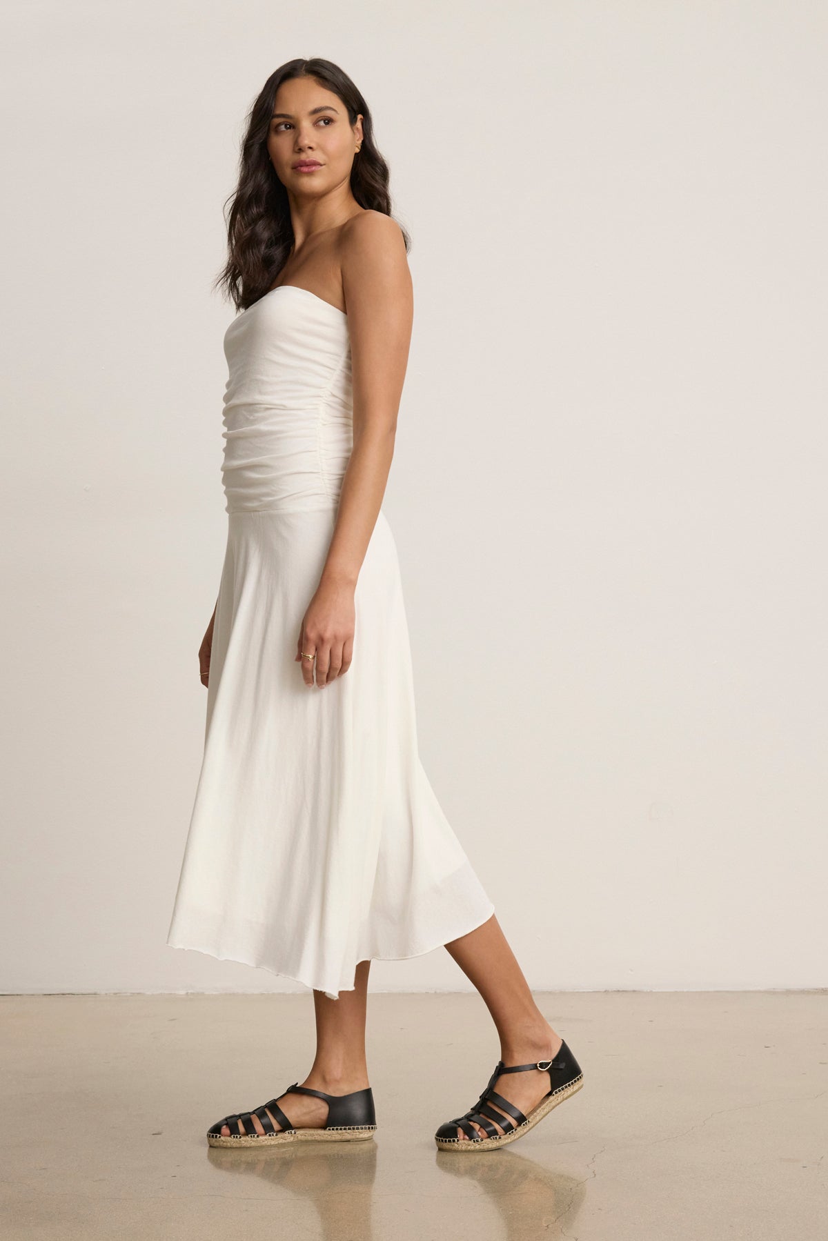 A woman stands sideways against a plain background, wearing the strapless SONIA DRESS by Velvet by Graham & Spencer and black sandals, looking slightly over her shoulder.-39145533243585