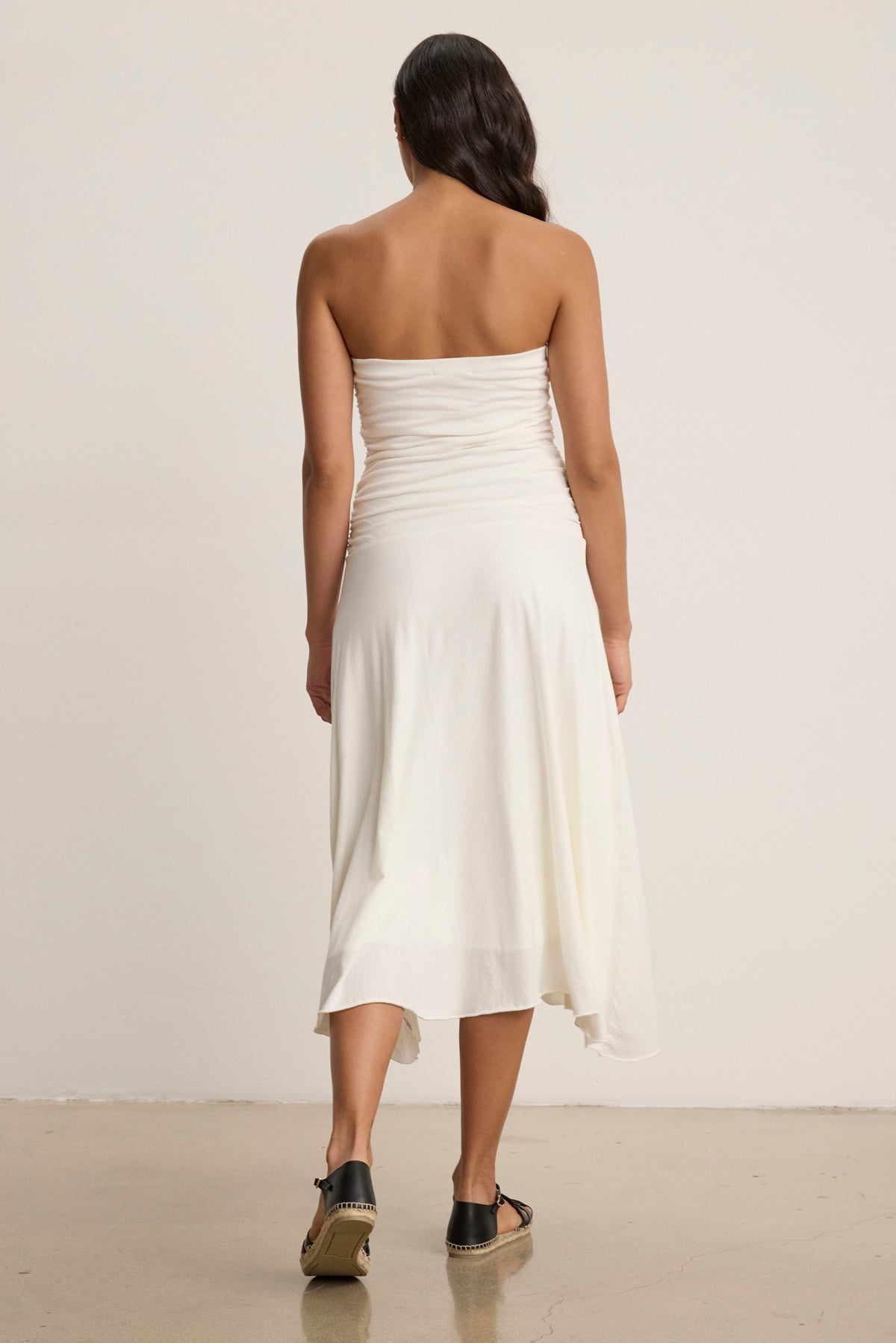 A woman stands with her back to the camera wearing the Velvet by Graham & Spencer SONIA DRESS, a strapless midi that creates a feminine silhouette, paired with black sandals in a plain indoor setting.-39145533210817