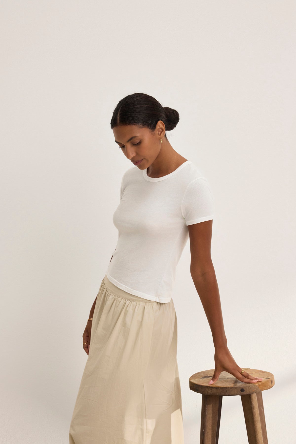 Wearing the WHITNEY TEE by Velvet by Graham & Spencer, a person in a beige skirt exudes classic style beside a wooden stool, gazing down against a plain background.-38928815390913