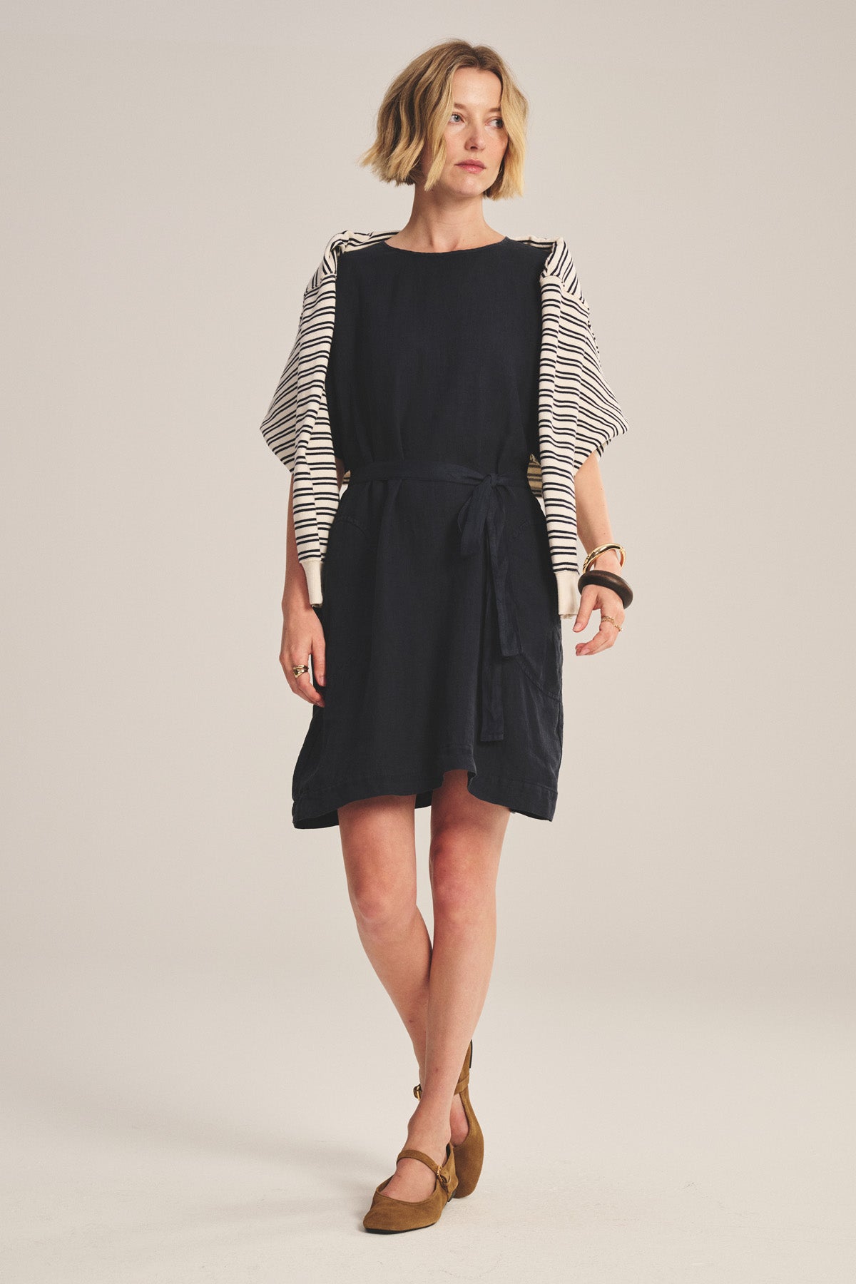 A woman wears the VIVIAN LINEN DRESS by Velvet by Graham & Spencer, a loose-fitting black mini dress with dolman sleeves and a detachable belt. She has short blond hair and brown flats.-41297003937985