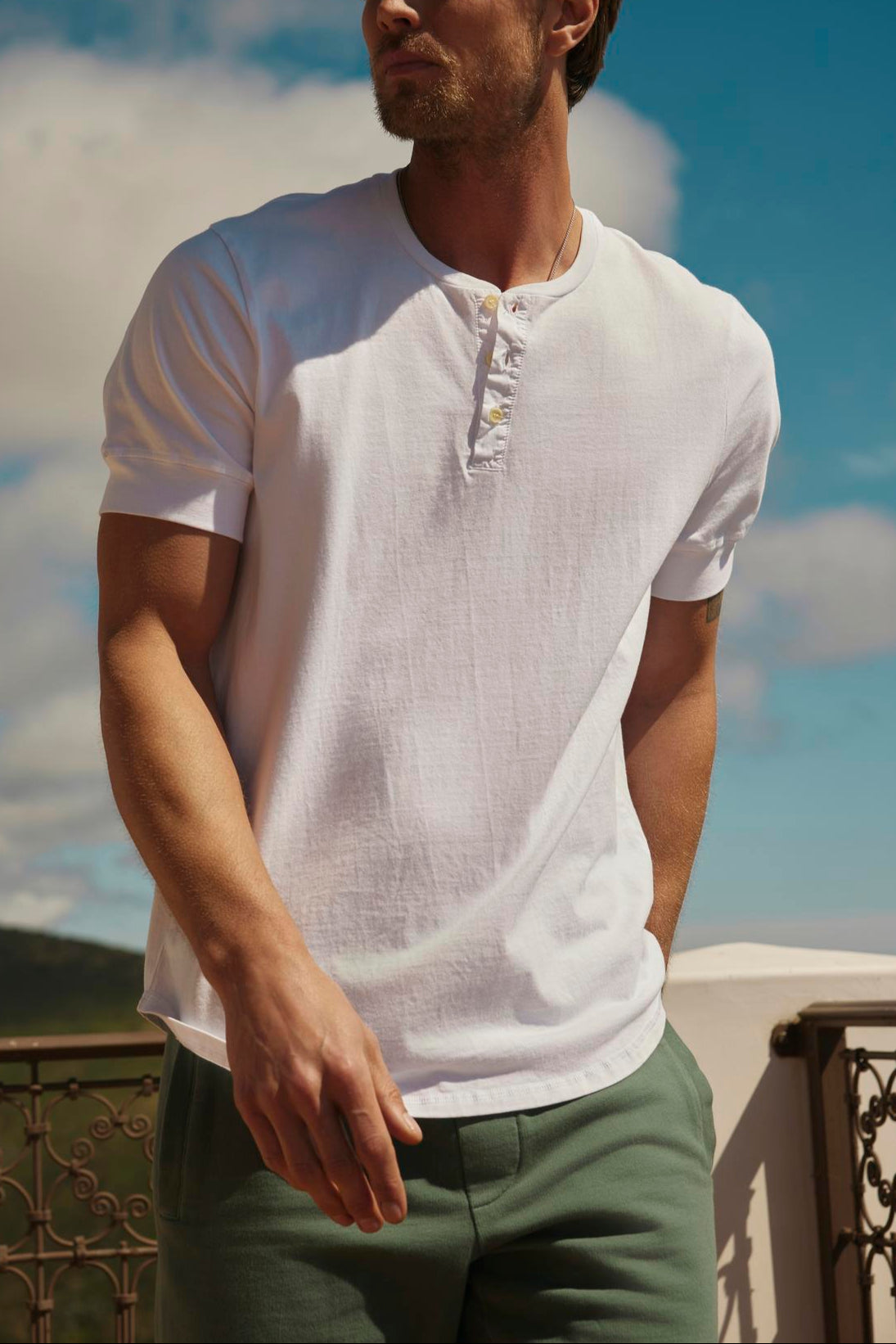 A man in a white Velvet by Graham & Spencer DEON HENLEY shirt with scooped hem and green trousers stands on a balcony, looking to the side with a backdrop of hills and sky.-36753582817473