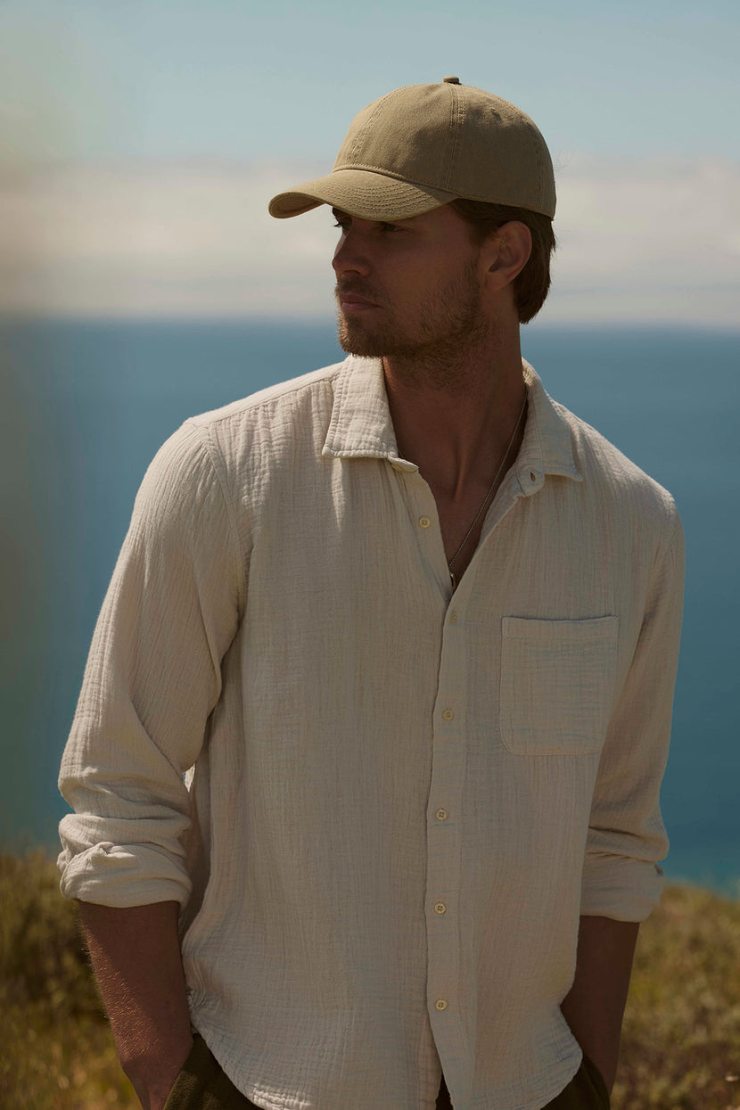 A man wearing a Velvet by Graham & Spencer ELTON COTTON GAUZE BUTTON-UP SHIRT and cap looking away thoughtfully against a coastal landscape backdrop.