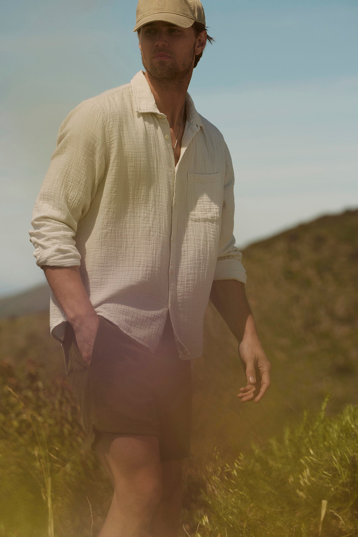   A man in an Elton Cotton Gauze button-up shirt and shorts stands in a sunny field, with a thoughtful expression and tousled hair, gazing into the distance. 