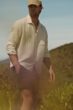 A man in an Elton Cotton Gauze button-up shirt and shorts stands in a sunny field, with a thoughtful expression and tousled hair, gazing into the distance.