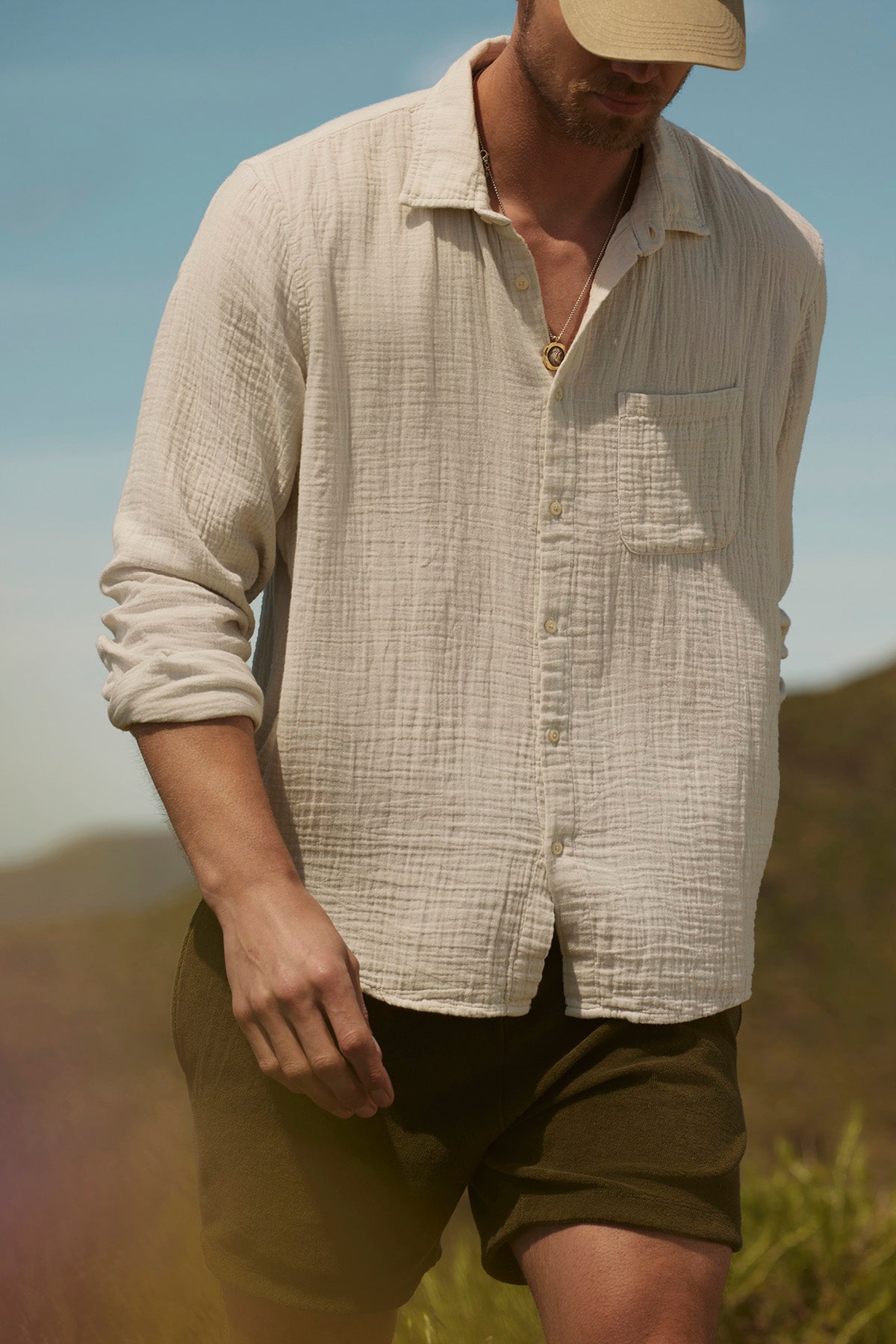 A man in a cream SALEM SHORT button-up shirt by Velvet by Graham & Spencer and a cap strolls outdoors against a blurred natural backdrop. The slash pockets on his shirt add casual elegance to his relaxed look.-38892260524225