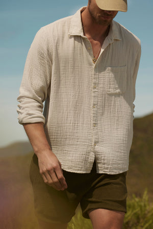 A man in a cream SALEM SHORT button-up shirt by Velvet by Graham & Spencer and a cap strolls outdoors against a blurred natural backdrop. The slash pockets on his shirt add casual elegance to his relaxed look.