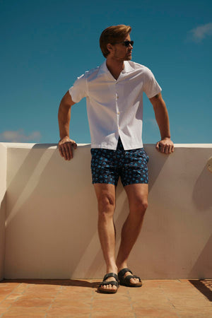 Man in sunglasses, short-sleeve white shirt, and Velvet by Graham & Spencer RICARDO SWIM SHORT standing on a sunny terrace, looking to the side.
