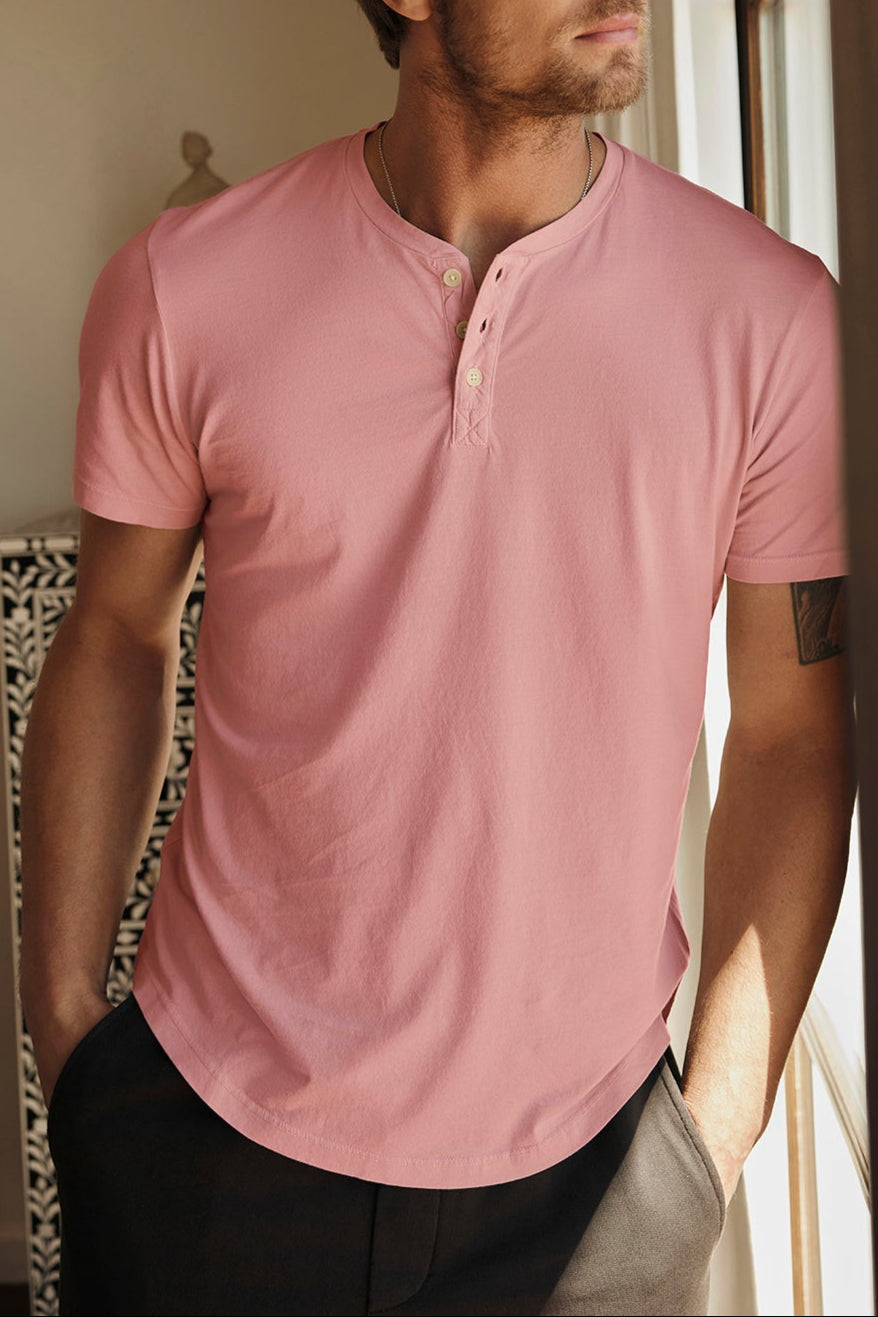 A man with light brown hair and facial hair stands by a window, wearing the FULTON HENLEY in pink by Velvet by Graham & Spencer, made from lightweight cotton jersey, along with dark pants, looking outwards.-37696180814017