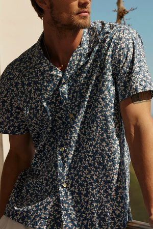 A man with light brown hair, wearing sunglasses and an IGGY BUTTON-UP SHIRT by Velvet by Graham & Spencer, featuring a blue floral design and a relaxed fit, leans against a railing with a scenic outdoor backdrop.