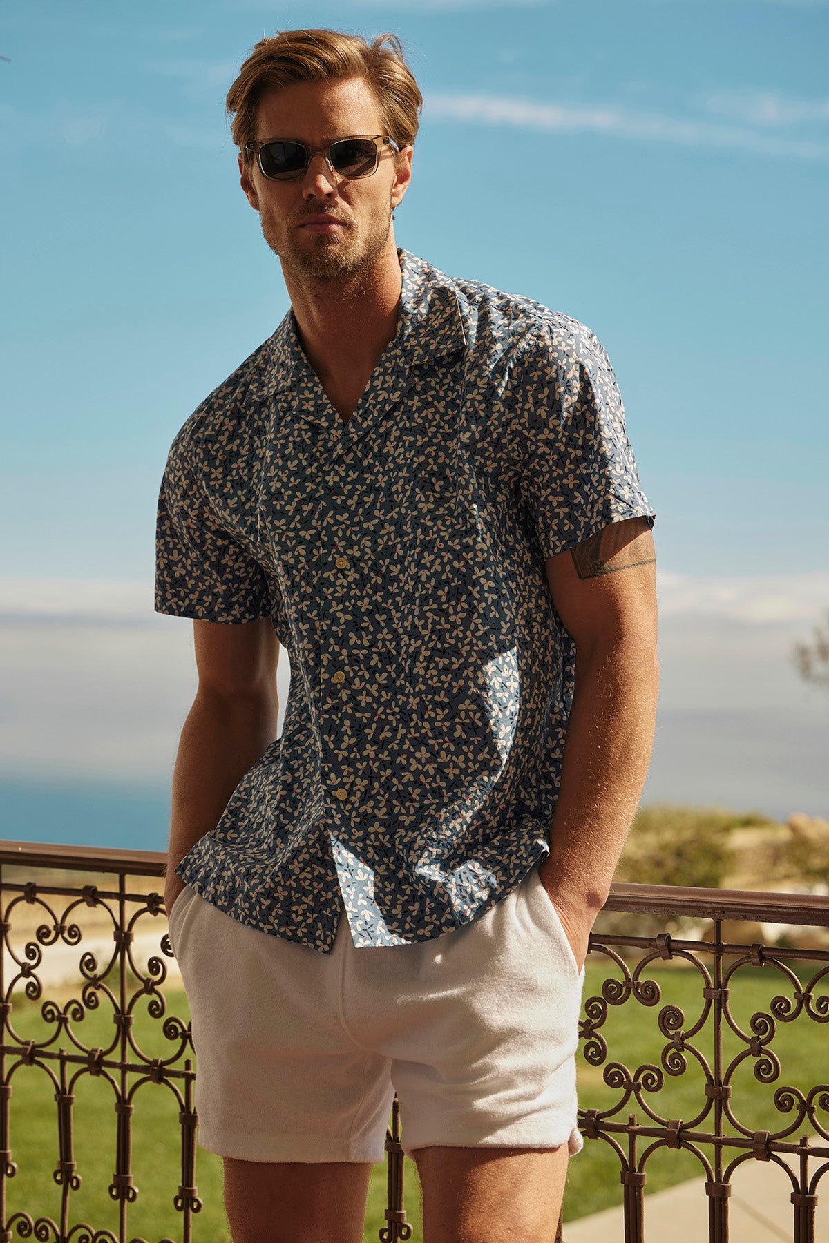 A man sporting sunglasses, a patterned short-sleeve shirt, and Velvet by Graham & Spencer's SALEM SHORTS, featuring a chic drawstring elastic waist, stands on an iron-railed balcony with a scenic view that enhances the laid-back vibe.