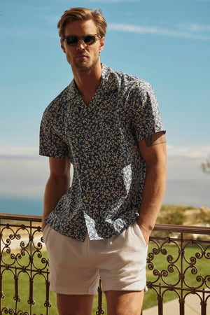 A man sporting sunglasses, a patterned short-sleeve shirt, and Velvet by Graham & Spencer's SALEM SHORTS, featuring a chic drawstring elastic waist, stands on an iron-railed balcony with a scenic view that enhances the laid-back vibe.