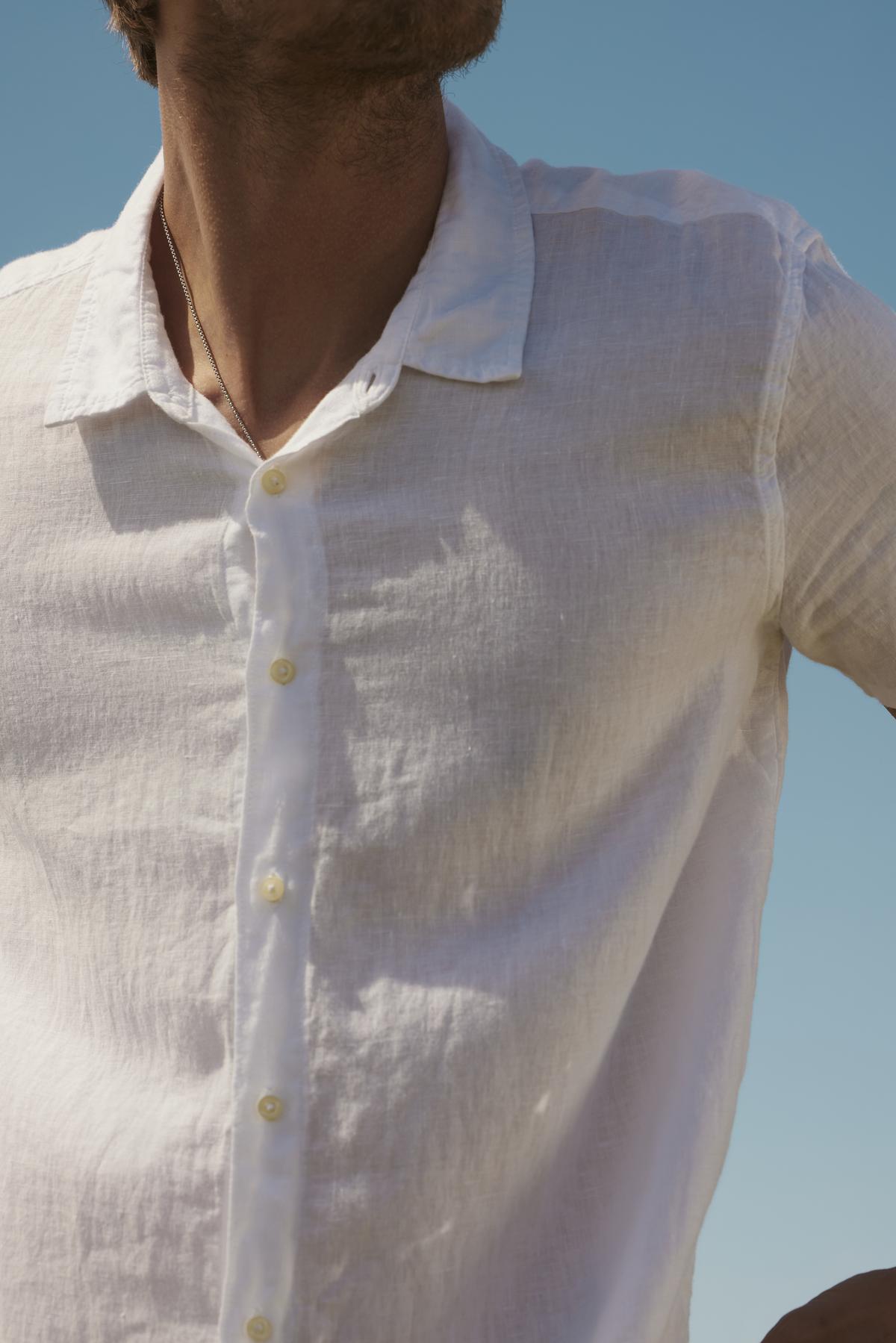 Close-up of a person wearing a Velvet by Graham & Spencer MACKIE LINEN BUTTON-UP SHIRT, focusing on the torso and unbuttoned collar. Clear blue sky in the background.