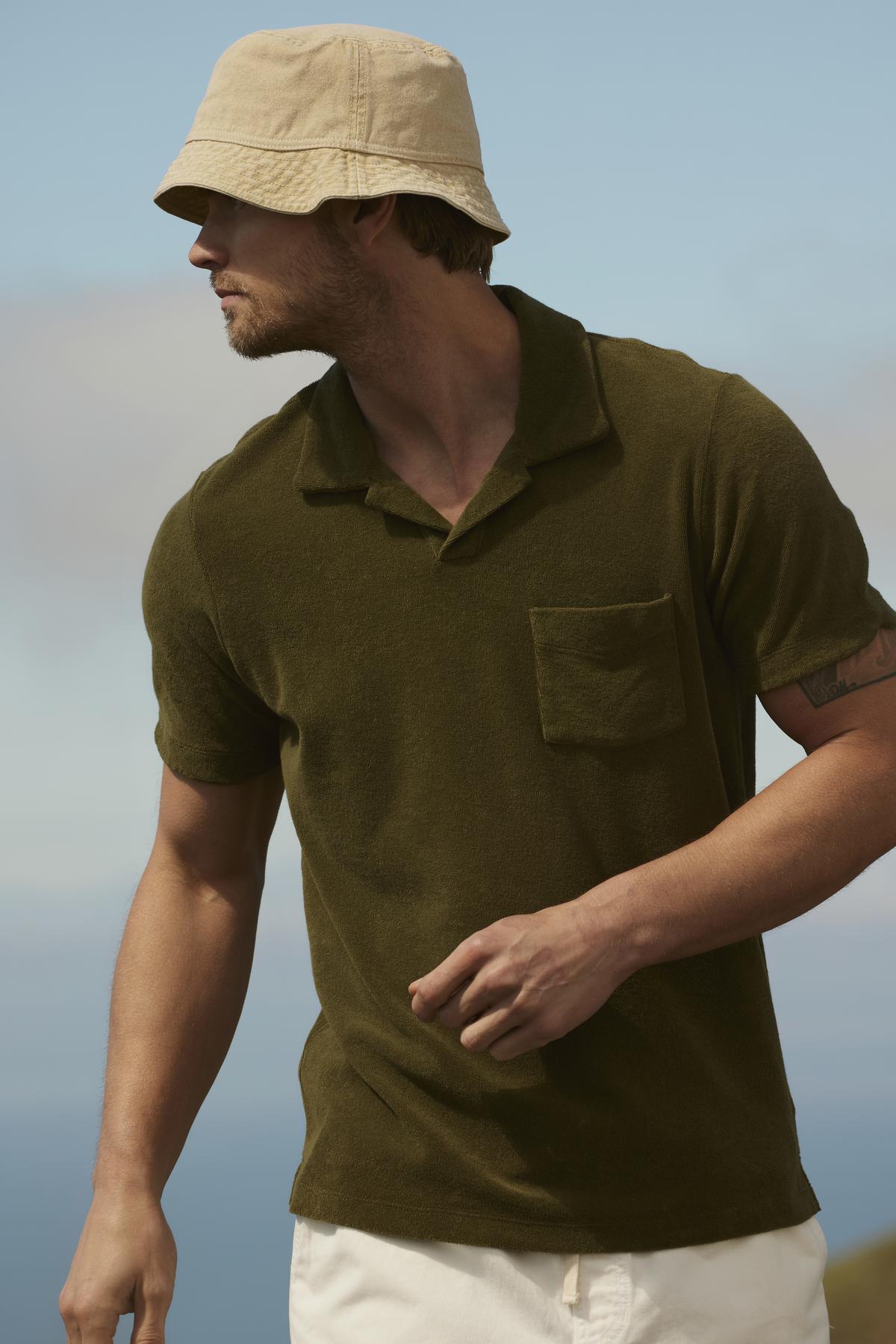 Man in a Velvet by Graham & Spencer Sergey polo shirt and beige hat, looking to the side, standing outdoors with a blurred landscape in the background.