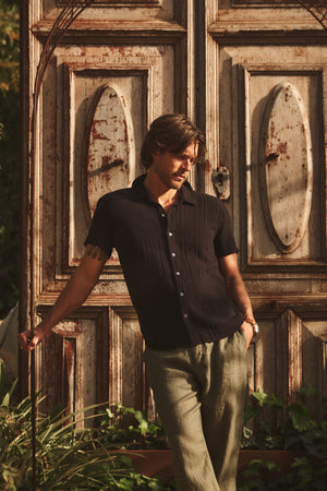 A man in the CHRISTIAN COTTON GAUZE BUTTON-UP SHIRT by Velvet by Graham & Spencer and light pants stands before large wooden doors, leaning on a cane and looking to the side.