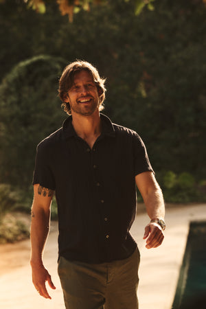 A man with mid-length hair and a beard smiles as he walks outdoors near greenery, wearing the CHRISTIAN COTTON GAUZE BUTTON-UP SHIRT by Velvet by Graham & Spencer with pants.