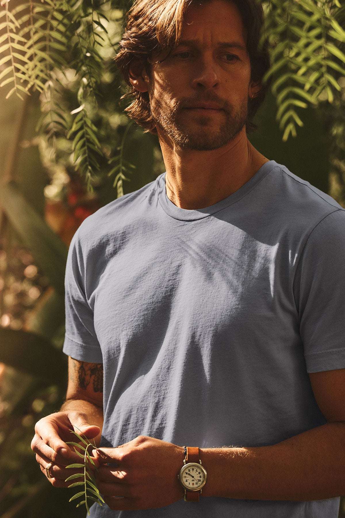 A brown-haired, bearded man stands outdoors among green plants, wearing the light blue HOWARD TEE by Velvet by Graham & Spencer and a wristwatch, gazing to the side with a vintage vibe.-40117592260801