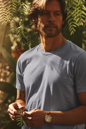 A brown-haired, bearded man stands outdoors among green plants, wearing the light blue HOWARD TEE by Velvet by Graham & Spencer and a wristwatch, gazing to the side with a vintage vibe.